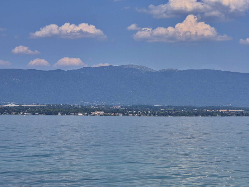 La vue en direction du sommet de la Dôle. Au milieu, au bord du rivage, le château de Coppet. La vue en direction du sommet de la Dôle. Au milieu, au bord du rivage, le château de Coppet. Plage des Courbes. Anières, lac Léman, canton de Genève.