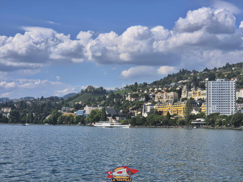 Un bateau en approche du débarcadère sur la droite. "Au-dessus" du bateau, le 2M2C, le Montreux Music & Convention Centre. Au milieu de l'image, le château du Châtelard. Un bateau en approche du débarcadère sur la droite. "Au-dessus" du bateau, le 2M2C, le Montreux Music & Convention Centre. Au milieu de l'image, le château du Châtelard.