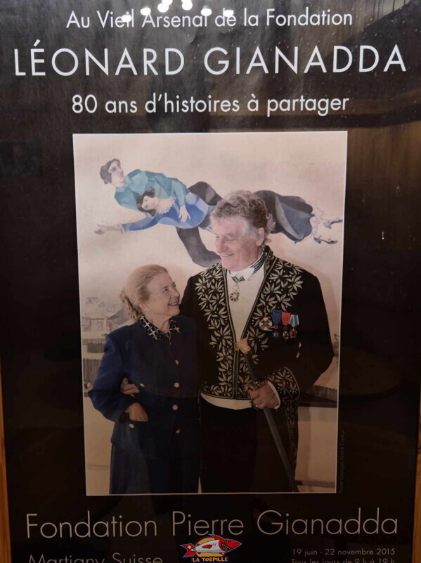 Une image montre l'affiche officielle de l'exposition rétrospective « Léonard Gianadda – 80 ans d'histoires à partager », qui s'est tenue au Vieil Arsenal de la Fondation Pierre Gianadda du 16 juin au 22 novembre 2015. La photo, en compagnie d'Anette, sa femme décédée en 2011, a été prise en décembre 2003 à Paris, à l'occasion de l'installation officielle de Léonard Gianadda à l'Académie des Beaux-Arts.